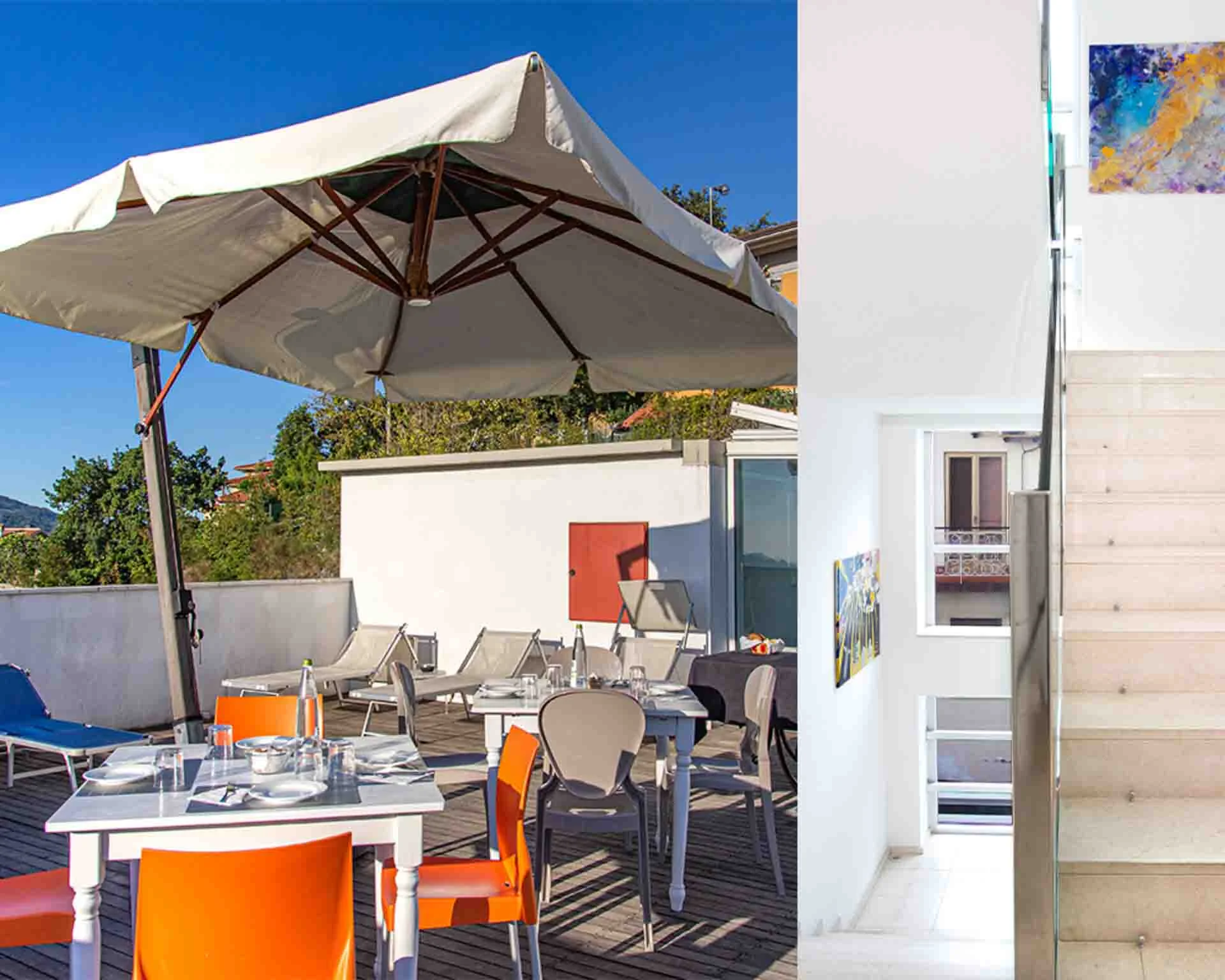 On the left or frame there is a roof top seating area with outdoor tables and orange chairs, on the left there is a stairwell with white marble steps and images hanging on the walls