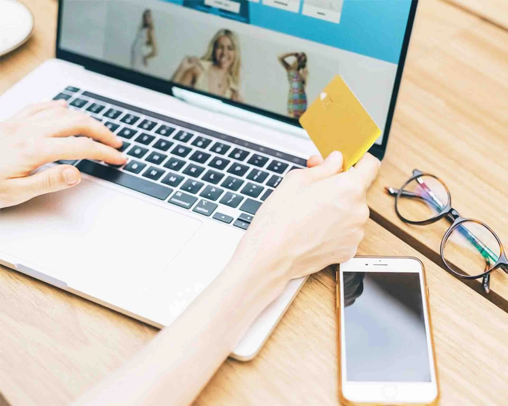 A laptop with an e-commerce web page being displayed and the person using the computer holding a yellow credit card
